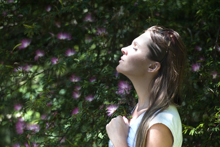 A woman enjoying a serene moment in a sunlit garden, surrounded by vibrant flowers and taking a deep breath.