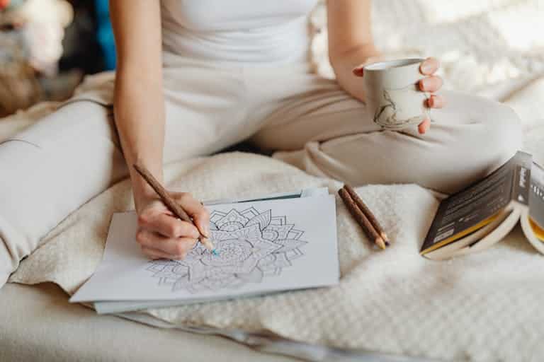 Artizen therapy blog. A woman enjoying a cozy moment with a coloring book and coffee in a bright, relaxed setting.